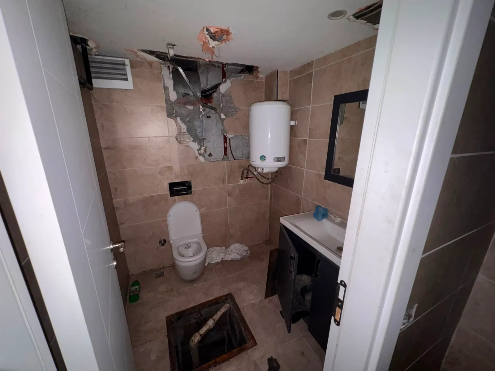 Kahverengi karolarla kaplı ıslak hacimde lavabo, ayna, termosifon ve tuvalet birlikte görülüyor.